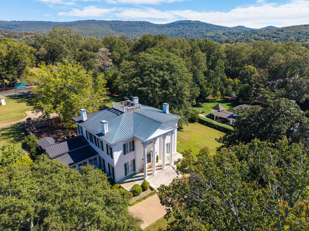 downtown-huntsville-mansion Aerial view of a white columned home surrounded by mature trees, with wooded hills in the distance.